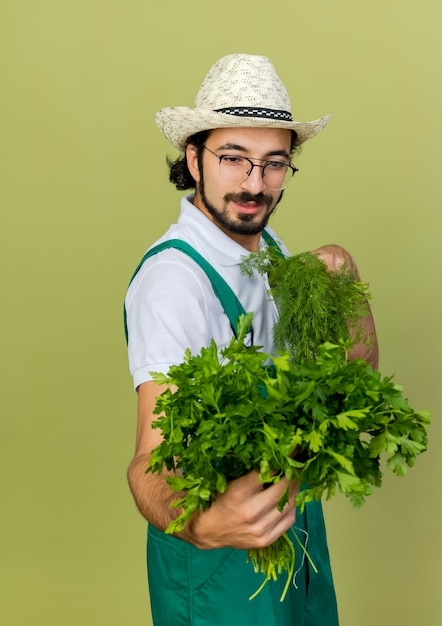 Pleased male gardener in optical glasses wearing gardening hat holds fennel and coriander