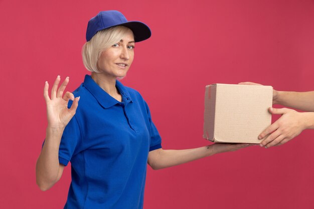 Pleased middle-aged blonde delivery woman in blue uniform and cap giving cardboard box to client doing ok sign 