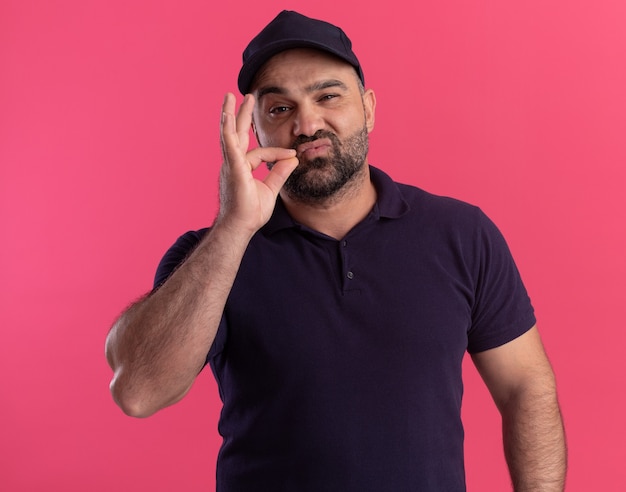 Pleased middle-aged delivery man in uniform and cap showing delicious gesture isolated on pink wall