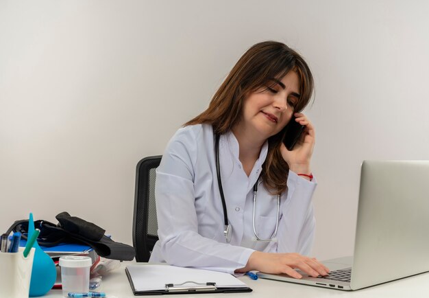 Pleased middle-aged female doctor wearing medical robe and stethoscope sitting at desk with medical tools clipboard and laptop talking on phone looking down using laptop isolated