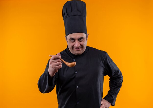 Pleased middle-aged male cook in chef uniform holding spoon on yellow wall