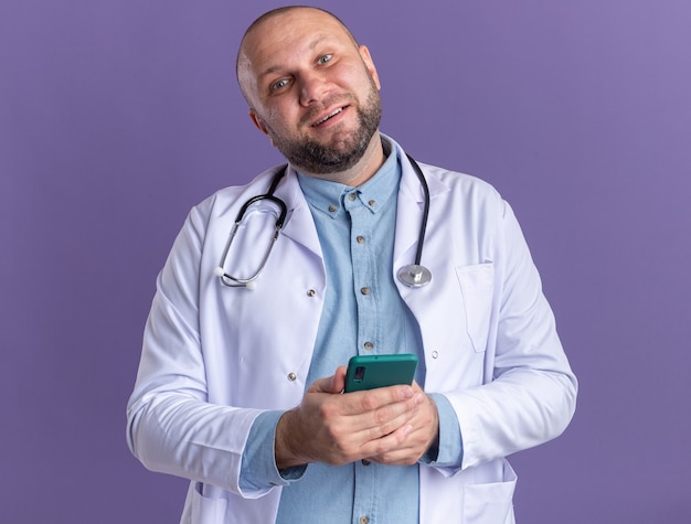 Free Photo pleased middle-aged male doctor wearing medical robe and stethoscope holding mobile phone  isolated on purple wall