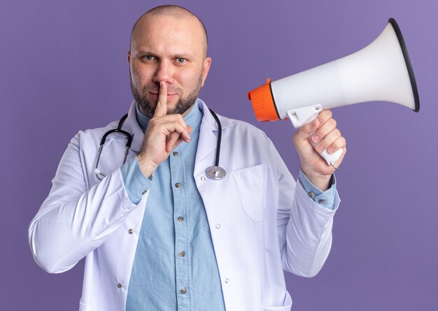 Pleased middle-aged male doctor wearing medical robe and stethoscope holding speaker  doing silence gesture isolated on purple wall