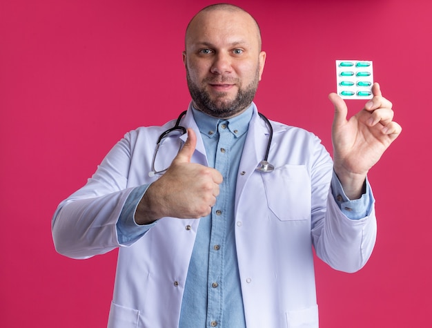 Pleased middle-aged male doctor wearing medical robe and stethoscope showing pack of medical capsules to front looking at front showing thumb up isolated on pink wall