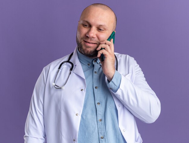 Pleased middle-aged male doctor wearing medical robe and stethoscope talking on phone looking down isolated on purple wall