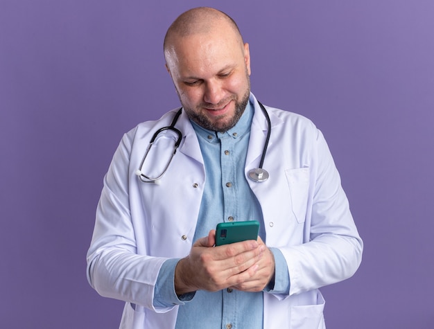 Free Photo pleased middle-aged male doctor wearing medical robe and stethoscope using his mobile phone isolated on purple wall