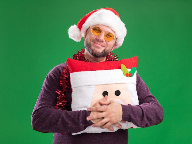 Free Photo pleased middle-aged man wearing santa hat and tinsel garland around neck with glasses holding santa claus pillow  isolated on green wall