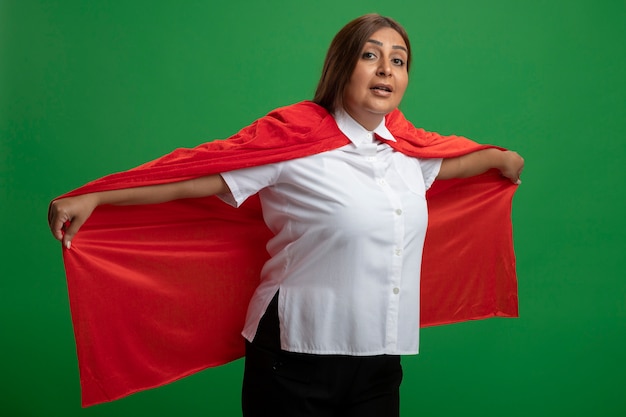 Pleased middle-aged superhero female holding cloak isolated on green background