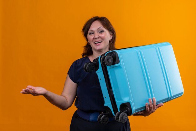 Pleased middle-aged traveler woman holding suitcase and points with hand to side on isolated orange background