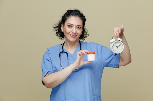 Pleased middleaged female doctor wearing uniform and stethoscope around neck looking at camera showing tablet container and alarm clock isolated on olive background