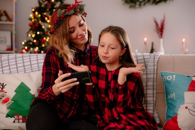 pleased mother holds phone and looks at confused daughter sitting on couch and enjoying christmas time at home