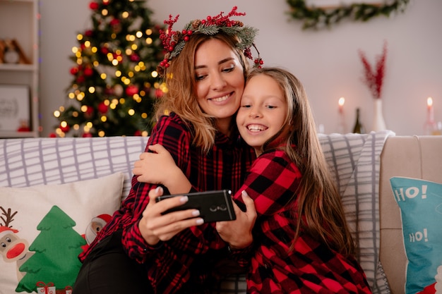 pleased mother with holly wreath and daughter watching something on phone sitting on couch and enjoying christmas time at home