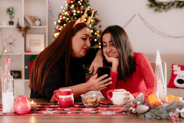 pleased mother with reindeer headband and daughter look at phone sitting at table enjoying the christmas time at home