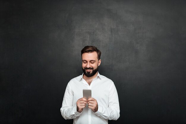 Pleased office worker in white shirt typing text message or scrolling feed in social network using cell phone over graphite