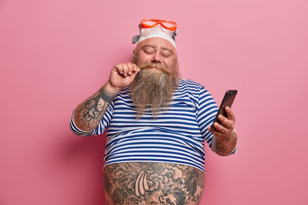 pleased overweight man stands with eyes closed touches mustache holds smartphone and checks newsfeed, wears swim goggles and striped sailor shirt enjoys good rest. Swimmer ready to dive