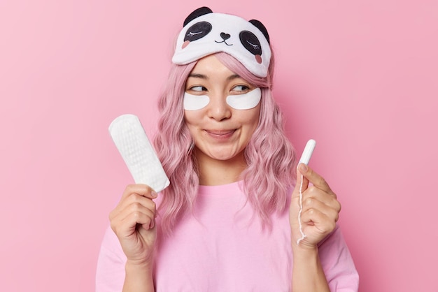 Free Photo pleased pink haired smiling asian woman has dreamy expression applies beauty patches under eyes for skin refreshment chooses best hygiene product holds sanitary napkin and tampon has menstruation