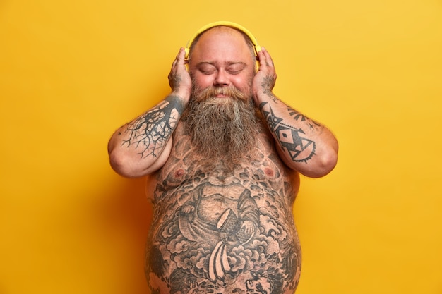 Pleased plump man listens music in headphones with enjoyment, closes eyes, stands naked, has tattooed body, fat sticking out tummy, thick beard, enjoys good sound, isolated on yellow wall