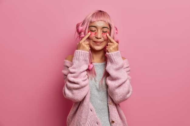 Free Photo pleased positive woman has long rosy hair, makes curly hairstyle with curlers, indicates at beauty patches, stands with closed eyes wears warm sweater