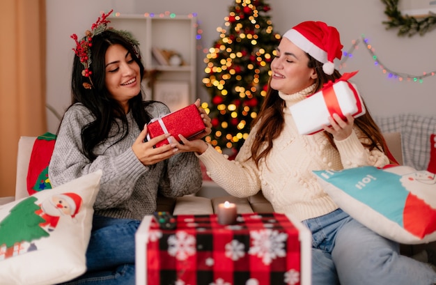 pleased pretty young girls with santa hat and holly wreath hold and look at gift boxes sitting on armchairs and enjoying christmas time at home