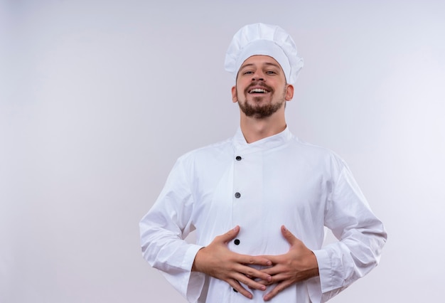 Pleased professional male chef cook in white uniform and cook hat holding his hands on his stomach standing over white background