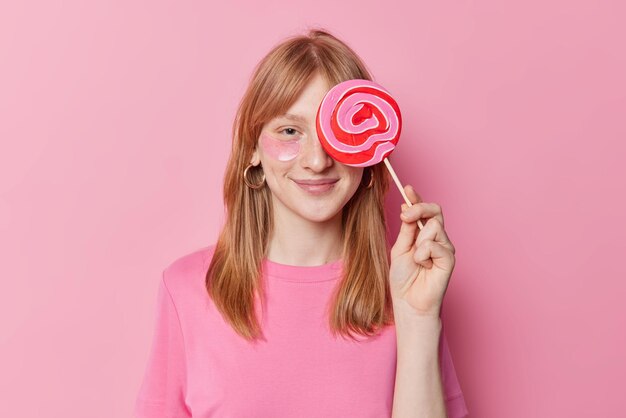 Pleased redhead girl covers eye with big delicious candy applies beauty patches wears casual t shirt poses against pink background has sweet tooth Children confectionary and beauty concept