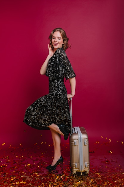 Pleased short-haired woman standing on one leg with suitcase and smiling with eyes closed