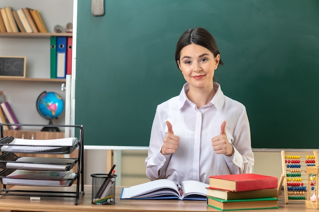 Pleased showing thumbs up young female teacher sitting at table with school tools in classroom