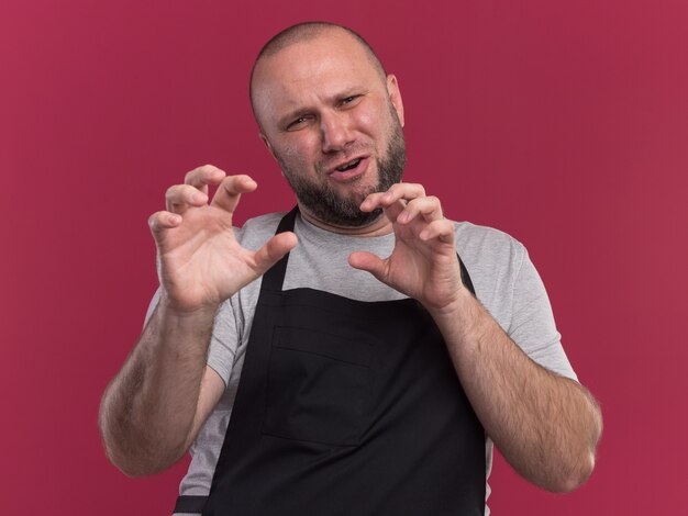 Pleased slavic middle-aged male barber in uniform showing tiger style gesture isolated on pink wall