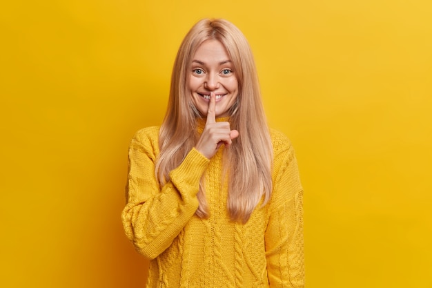 Pleased smiling blonde woman makes secret gesture, finger on lip, shushing