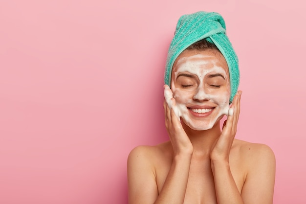 Pleased smiling woman washes face with cleansing gel, has soap on complexion, keeps eyes shut, wears wrapped towel on head, has naked body