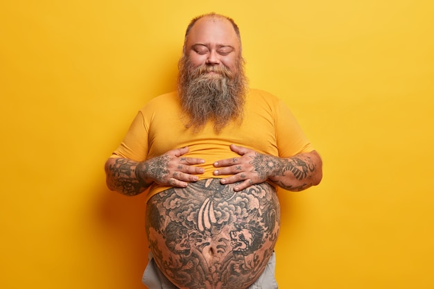 Free photo pleased stout man keeps hands on belly, feels satiety after eating delicious dinner, stands with eyes closed, doesnt care about figure, has hormonal imbalance, isolated on yellow wall.