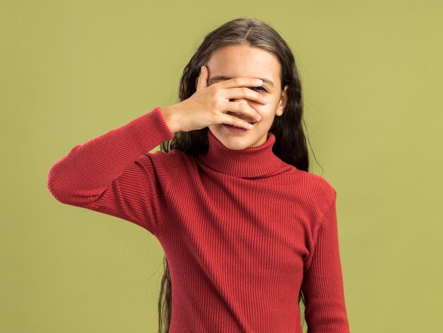 Pleased teenage girl looking at camera covering eyes with hand looking at camera through fingers isolated on olive green wall