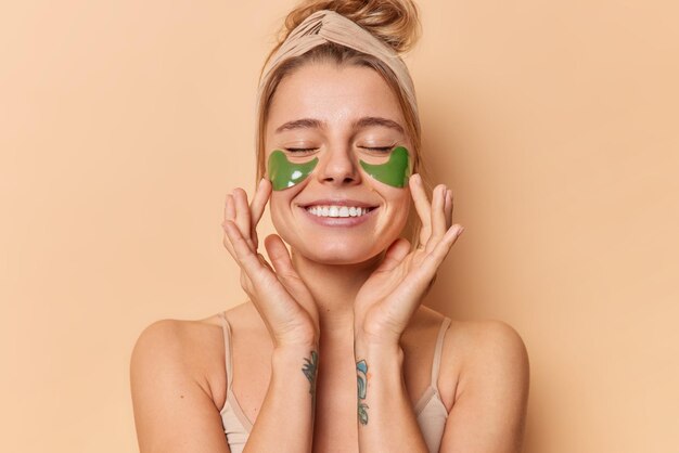 Pleased tender young woman applies green hydrogel patches under eyes to remove wrinkles wears headband has healthy clean skin white even teeth isolated over beige background Wellness concept