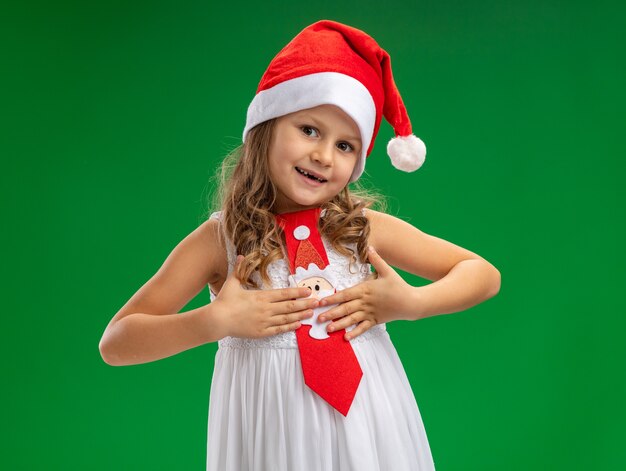 Pleased tilting head little girl wearing christmas hat with tie putting hands on herself isolated on green background