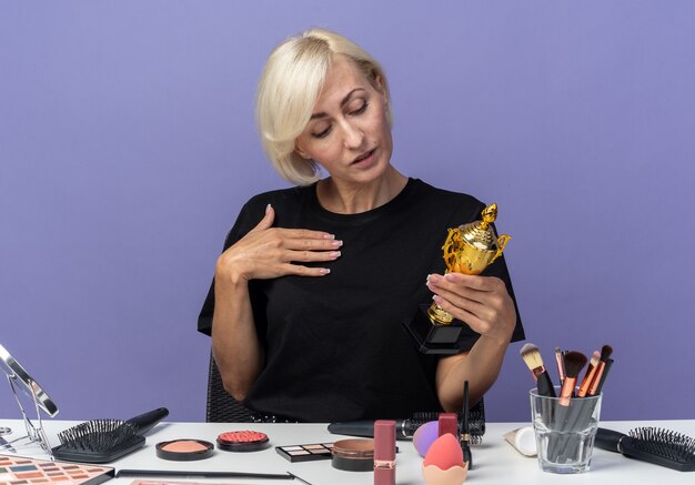 Pleased tilting head young beautiful girl sits at table with makeup tools holding winner cup isolated on blue wall