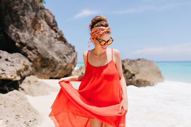 Pleased white female model in sunglasses chilling at beach with rocks. Cute caucasian girl playing with her red dress near ocean.