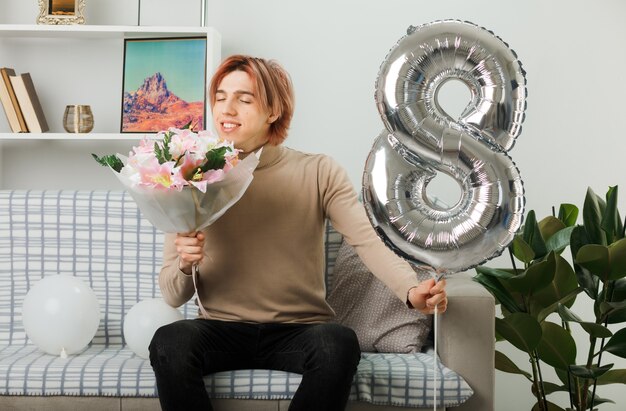 Pleased with closed eyes handsome guy on happy women day holding number eight balloon and sniffing bouquet in his hand sitting on sofa in living room