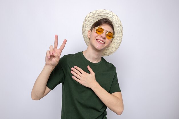 Pleased with closed eyes showing peace gesture young handsome guy wearing glasses with hat 