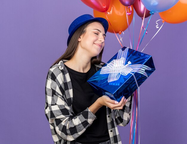 Pleased with closed eyes young beautiful girl wearing party hat holding balloons with gift box 