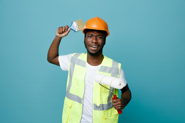 Pleased young african american builder in uniform holding roller brush with paint brush isolated on blue background