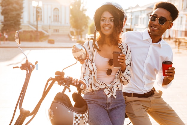 Pleased young african couple drinking coffee while standing near the modern motorbike on the street and looking away