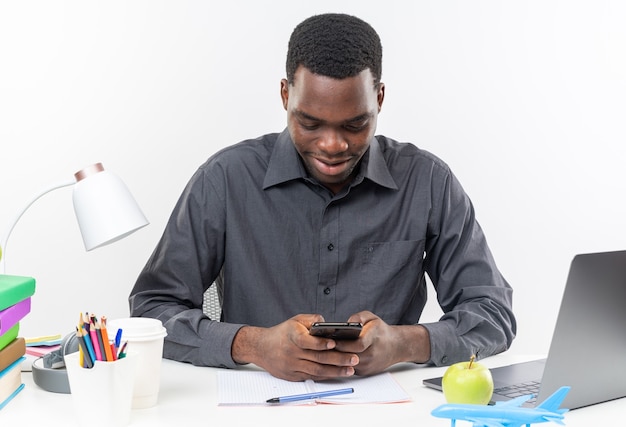 Pleased young afro-american student sitting at desk with school tools holding and looking at phone 