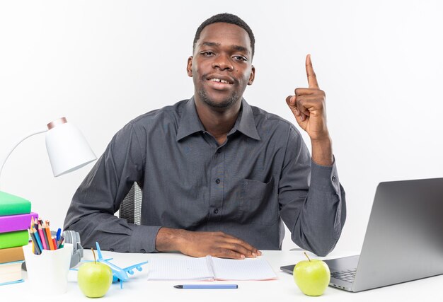 Pleased young afro-american student sitting at desk with school tools pointing up 