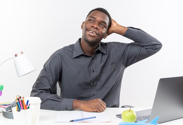 Pleased young afro-american student sitting at desk with school tools putting hand on his head and looking at side 