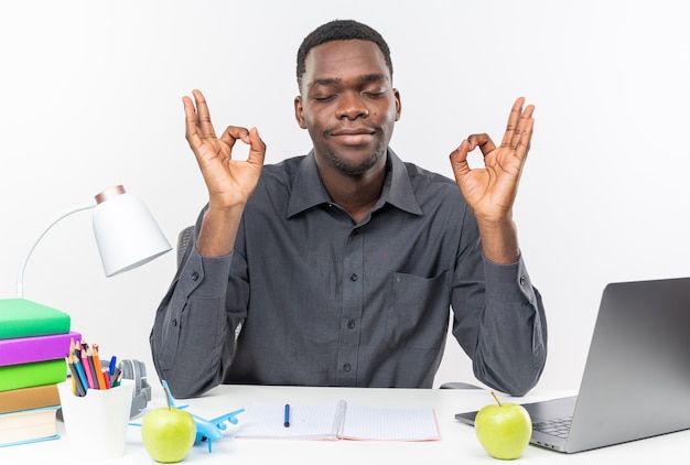 Pleased young afro-american student sitting with closed eyes at desk with school tools meditating isolated on white wall