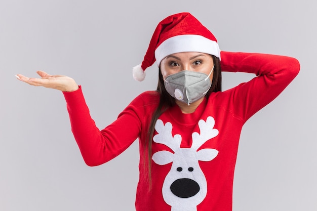 Free Photo pleased young asian girl wearing christmas hat with sweater and medical mask putting hand on head isolated on white wall