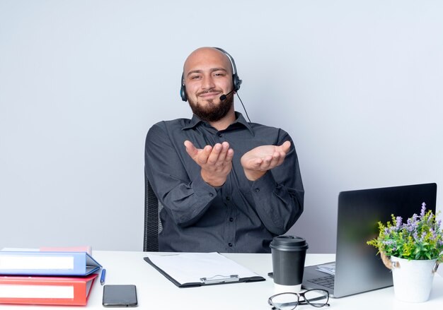 Pleased young bald call center man wearing headset sitting at desk with work tools showing empty hands at camera isolated on white background