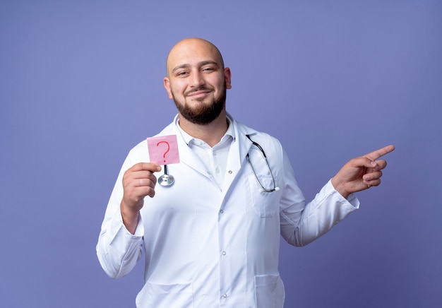 Pleased young bald male doctor wearing medical robe and stethoscope holding paper question mark and points at side