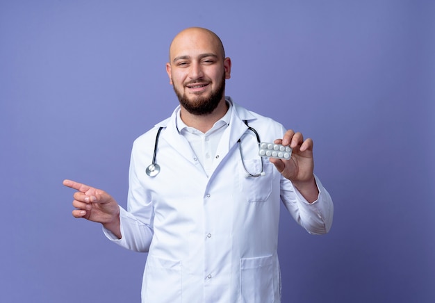 Pleased young bald male doctor wearing medical robe and stethoscope holding pills and points at side isolated on blue background with copy space