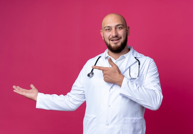 Pleased young bald male doctor wearing medical robe and stethoscope pretending holding something isolated on pink background with copy space